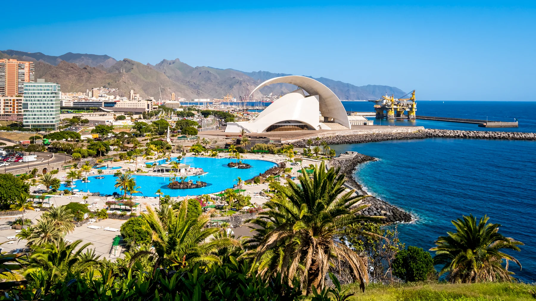 santa cruz de tenerife mit dem ikonischen konzertsaal auditorio de tenerife, der im sonnenlicht vor der kulisse des anaga-gebirges, einer bohrinsel und einem tropischen pool mit palmen im vordergrund glänzt.
